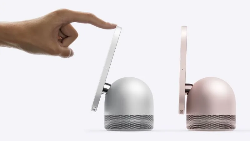 Two Apple HomeView displays in profile on their rounded speaker bases, in silver and rose pink, with a hand tilting one screen