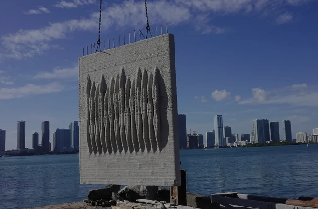 A large textured living seawall panel hanging from chains above the water with the Miami skyline across the bay under a blue sky