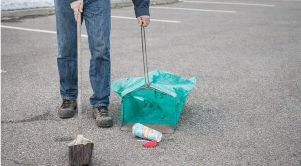 A worker in jeans holding a broom and a green litter collection bag in an empty parking lot, with a dropped cup on the asphalt