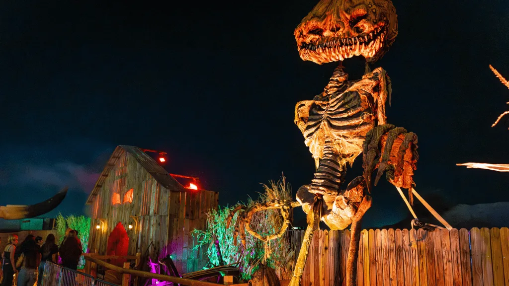 A giant pumpkin headed skeleton figure towering over guests at night at a Thirteenth Floor haunted attraction, with a haunted barn lit in red and green behind it