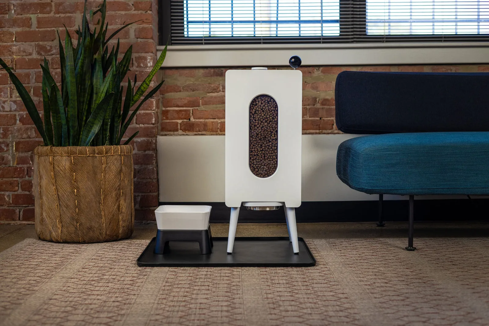 A white Houndsy kibble dispenser filled with dog food standing on a mat beside a water bowl, a plant, and a blue sofa in a brick walled room