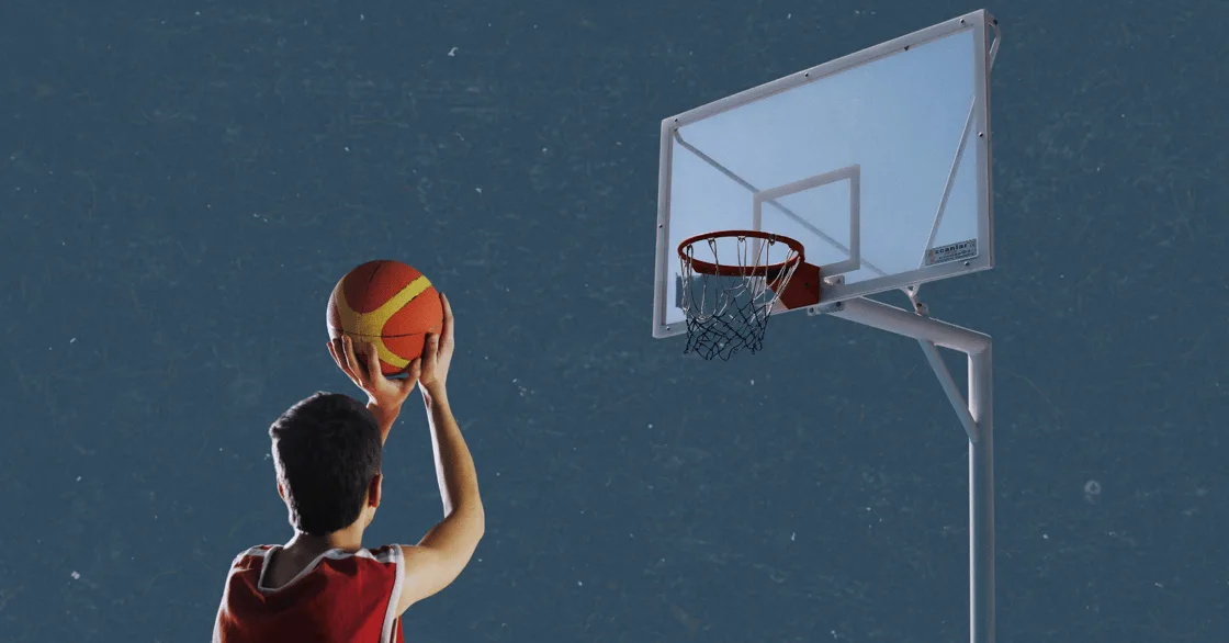 A young player in a red jersey shooting a basketball toward an outdoor hoop against a dark blue background