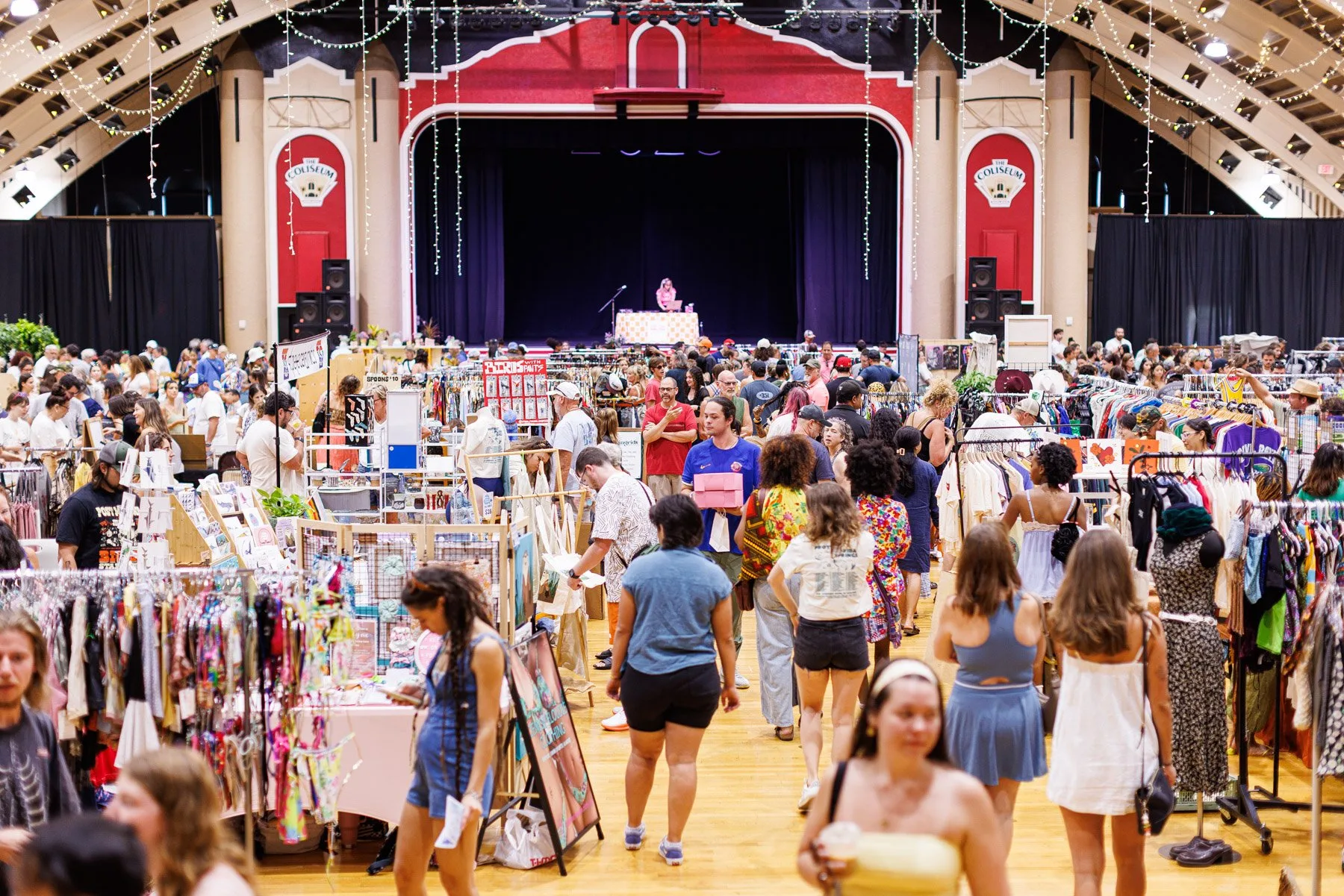 A crowded Indie Flea market hall with shoppers browsing clothing racks, art stalls, and maker booths under string lights in front of a stage
