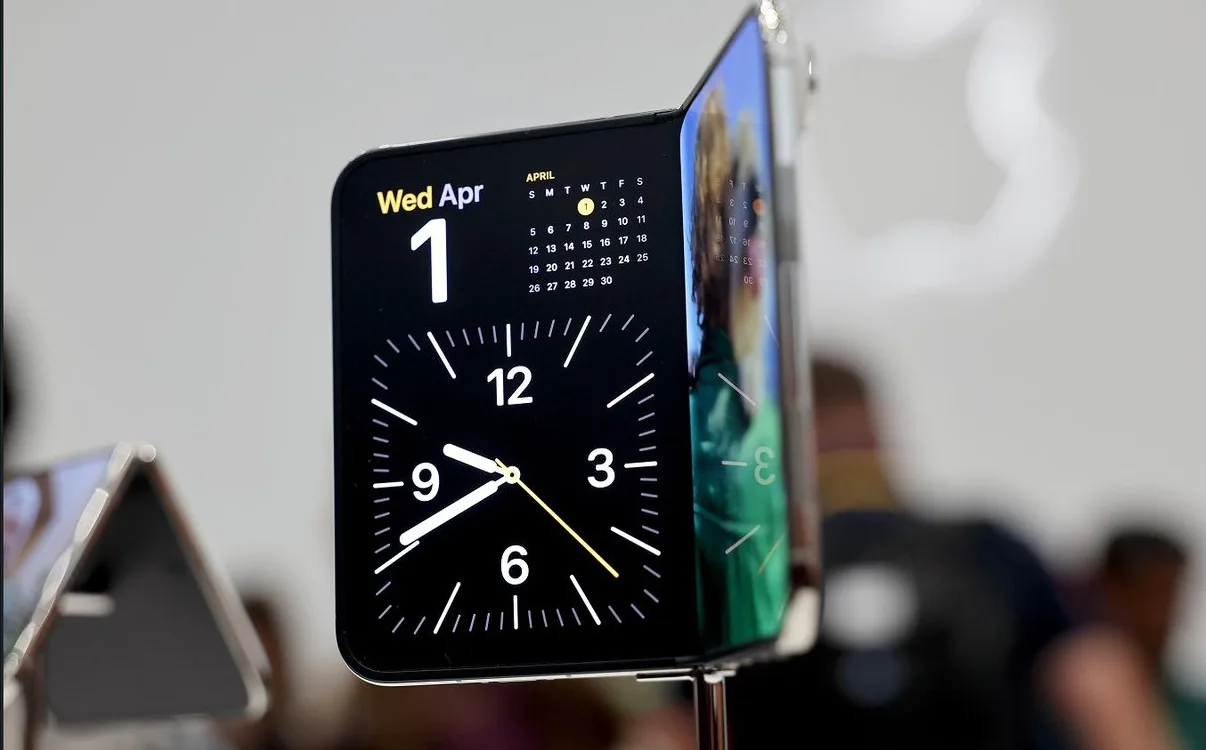 The folding iPhone Duo on display, half open, showing a large clock and April calendar on its inner screen with an Apple logo blurred in the background