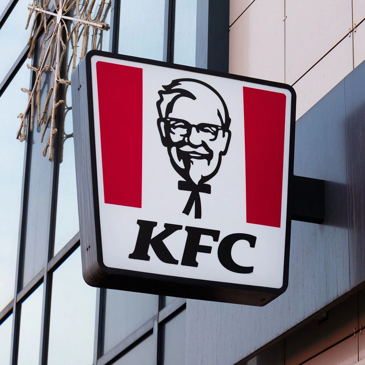 A red and white KFC sign with the Colonel Sanders portrait mounted on the glass front of a restaurant building