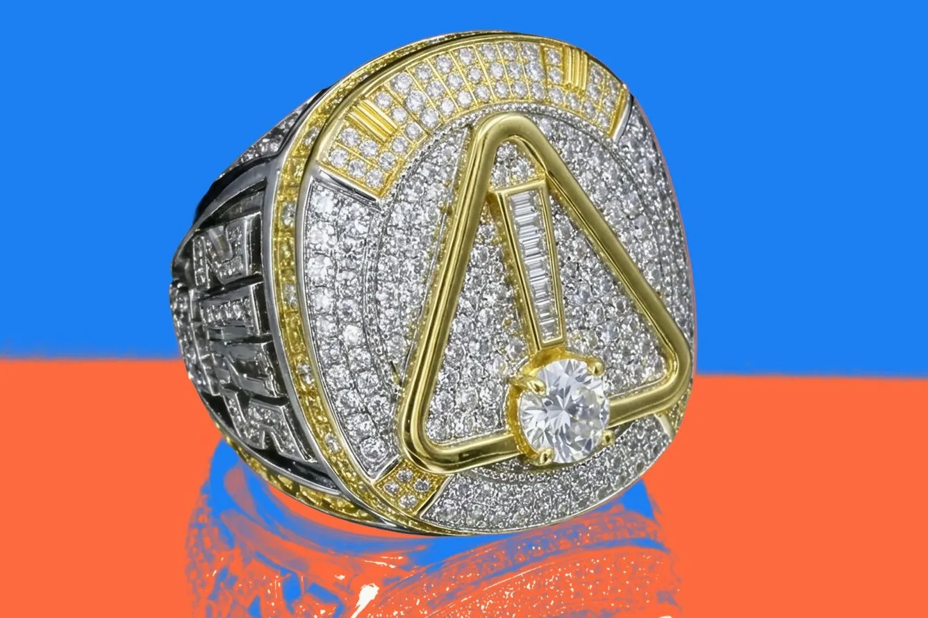 A diamond covered championship ring with a gold warning sign on its face, on a blue and orange background