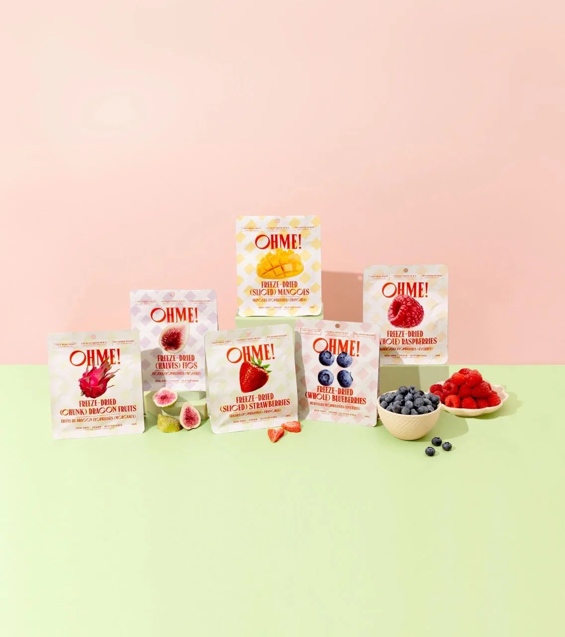 Six OHME pouches of freeze dried fruit, mango, fig, strawberry, blueberry, raspberry, and dragon fruit, arranged with bowls of fresh berries on a pink and green background