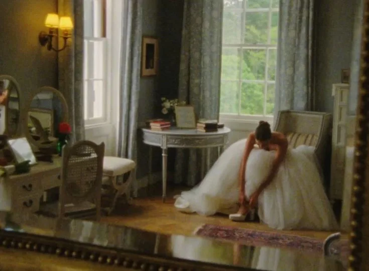 A bride in a white wedding dress putting on her shoe in a bright room with tall windows, captured in the soft candid style of a wedding home movie still