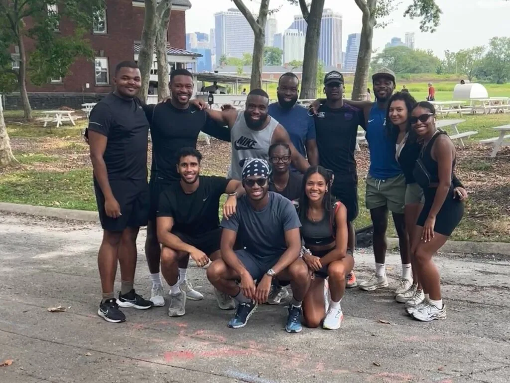 Members of the Peak and Pace run club posing together outdoors in running gear with the New York skyline and picnic tables behind them
