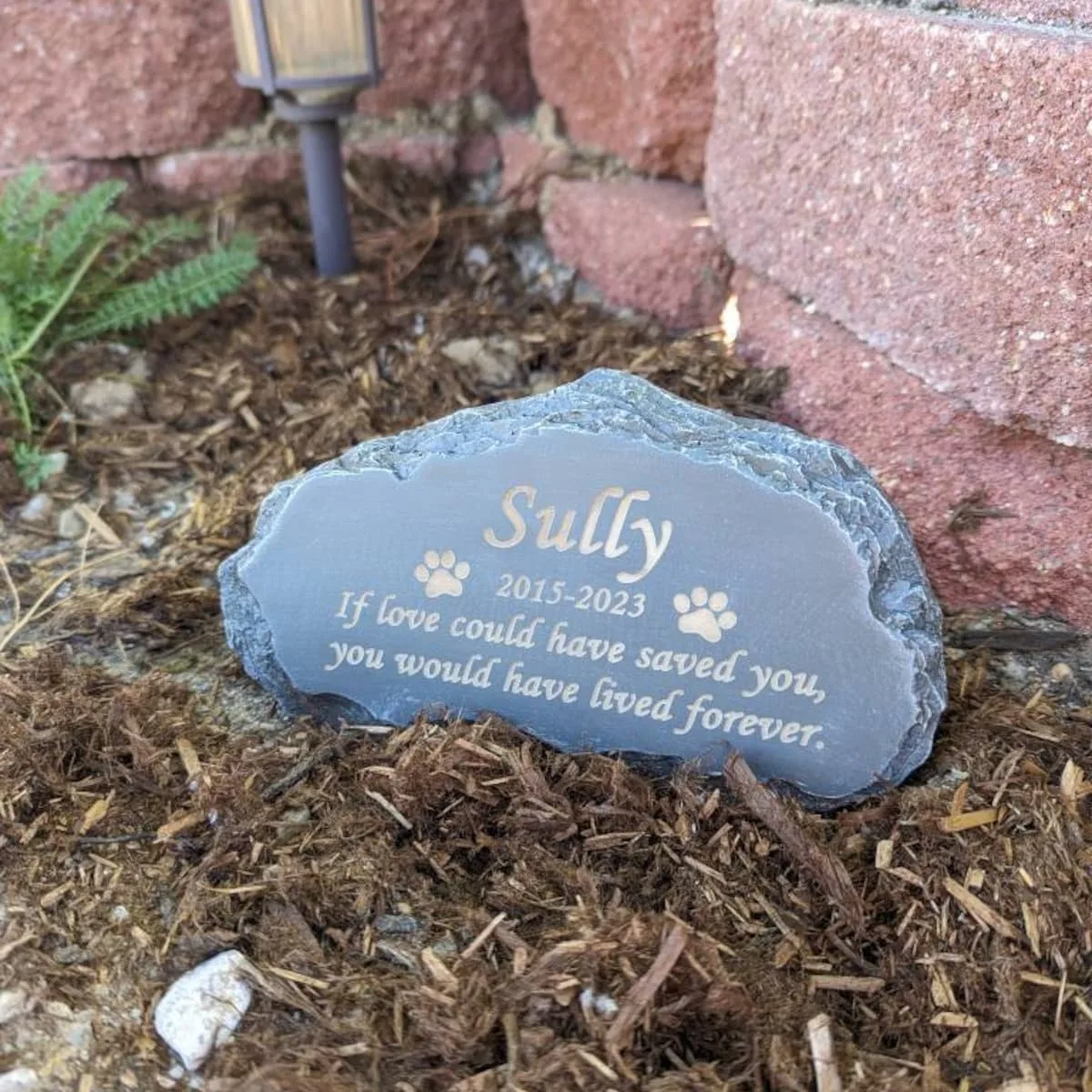 An engraved pet memorial stone set in mulch beside a brick wall, carrying a dogs name, the years 2015 to 2023, paw prints, and a remembrance message