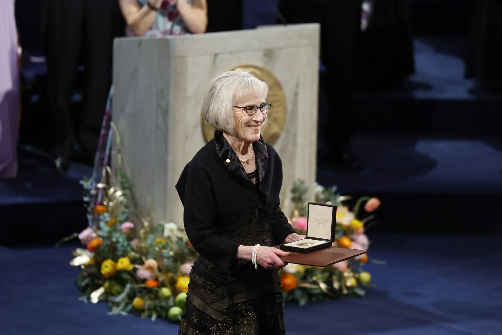 Economist Claudia Goldin smiling as she holds her Nobel Prize medal in an open case on a stage decorated with flowers