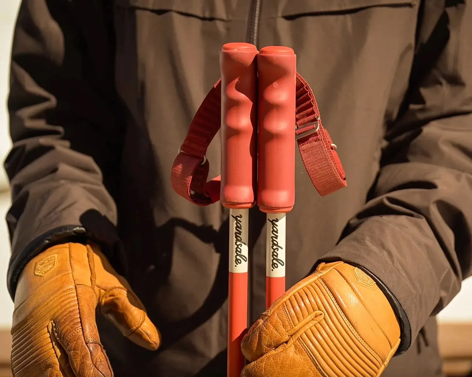 A skier in leather gloves holding two red Yardsale ski poles clicked together by their magnetic handles, with the Yardsale name printed on the shafts